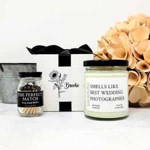 May include: A gift set featuring a white box with a black ribbon, a small glass jar of matches labeled THE PERFECT MATCH, and a glass jar candle labeled SMELLS LIKE BEST WEDDING PHOTOGRAPHER, NET WT 9OZ, HAND POURED IN TENNESSEE.