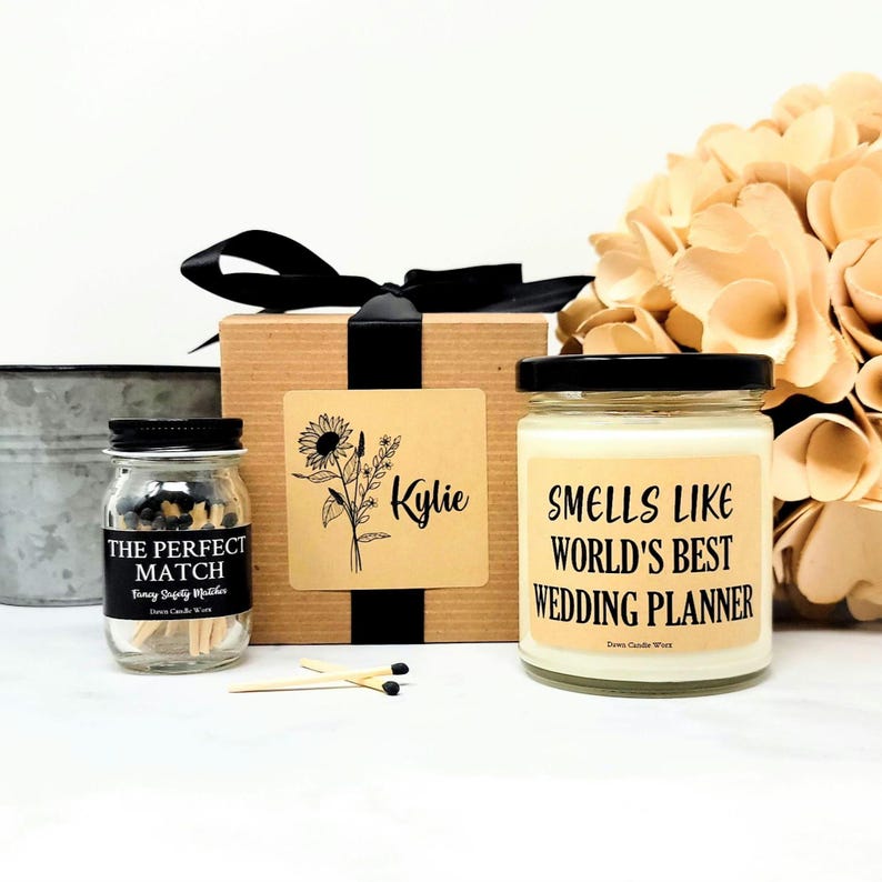 May include: A gift set featuring a candle, matches, and a decorative box. The candle jar has a black lid and a label that reads "SMELLS LIKE WORLD'S BEST WEDDING PLANNER." The match jar is labeled "THE PERFECT MATCH." The box has a sunflower design.