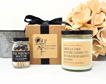 May include: A gift set with a candle, matches, and a box tied with a black ribbon. The candle has a label that reads "Smells Like You're Going to Be Grandparents Again!" The match jar reads "The Perfect Match."