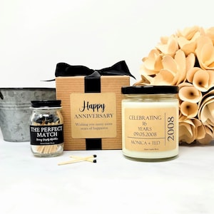 May include: A gift set featuring a brown cardboard box with a black ribbon, a small glass jar of matches labeled The Perfect Match, and a glass candle jar labeled Celebrating 16 Years 09.05.2008 Monica + Ted 2008. Two loose matches lie in front.