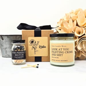 May include: A gift set featuring a candle, matches, and a decorative box. The candle has a black lid and a label that reads "LOOK AT YOU FIGHTING CRIME AND SHIT." The match jar is labeled "THE PERFECT MATCH." The box is adorned with a sunflower.