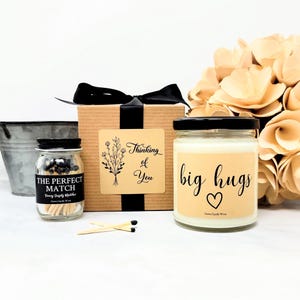 May include: A gift set featuring a brown cardboard box with a black ribbon, a small glass jar of matches labeled The Perfect Match, and a glass candle jar with a cream-colored label reading big hugs with a heart icon and two loose matches.