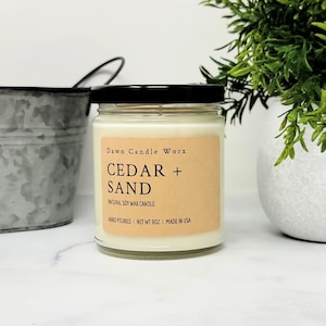 May include: A clear glass jar candle with a black lid and a light brown rectangular label. The label text reads Dawn Candle Worx, CEDAR + SAND, NATURAL SOY WAX CANDLE, HAND POURED, NET WT 9OZ, MADE IN USA. The candle contains off-white wax.