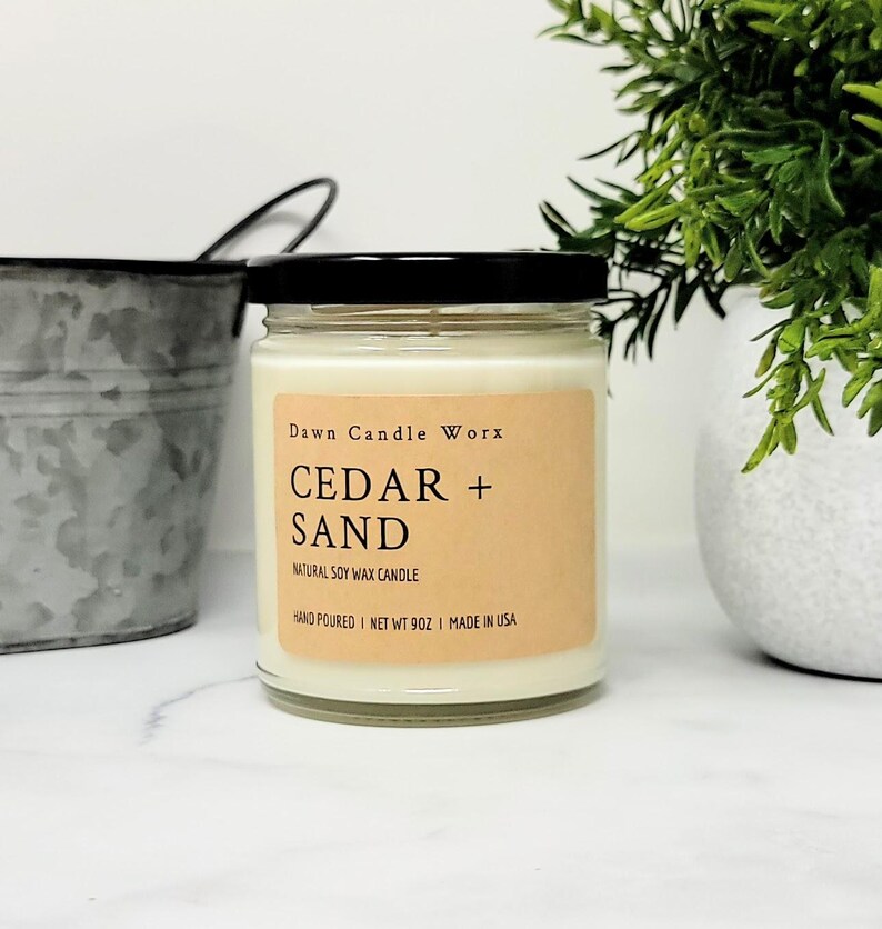 May include: A clear glass jar candle with a black lid and a light brown rectangular label. The label text reads Dawn Candle Worx, CEDAR + SAND, NATURAL SOY WAX CANDLE, HAND POURED, NET WT 9OZ, MADE IN USA. The candle contains off-white wax.