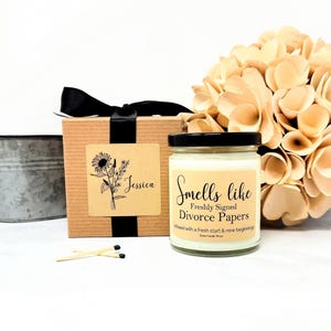 K&ouml;nnte beinhalten: Ein Glaskerzenglas mit schwarzem Deckel und einem Etikett mit der Aufschrift Smells like Freshly Signed Divorce Papers, neben einer braunen Geschenkbox mit schwarzer Schleife und einem Etikett mit Blumenillustration und dem Namen Jessica sowie zwei Streichh&ouml;lzern.