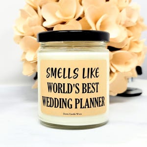 May include: A glass jar candle with a black lid and a label that reads "Smells Like World's Best Wedding Planner." The candle has a light beige label with black text. The background features a bouquet of light-colored flowers.