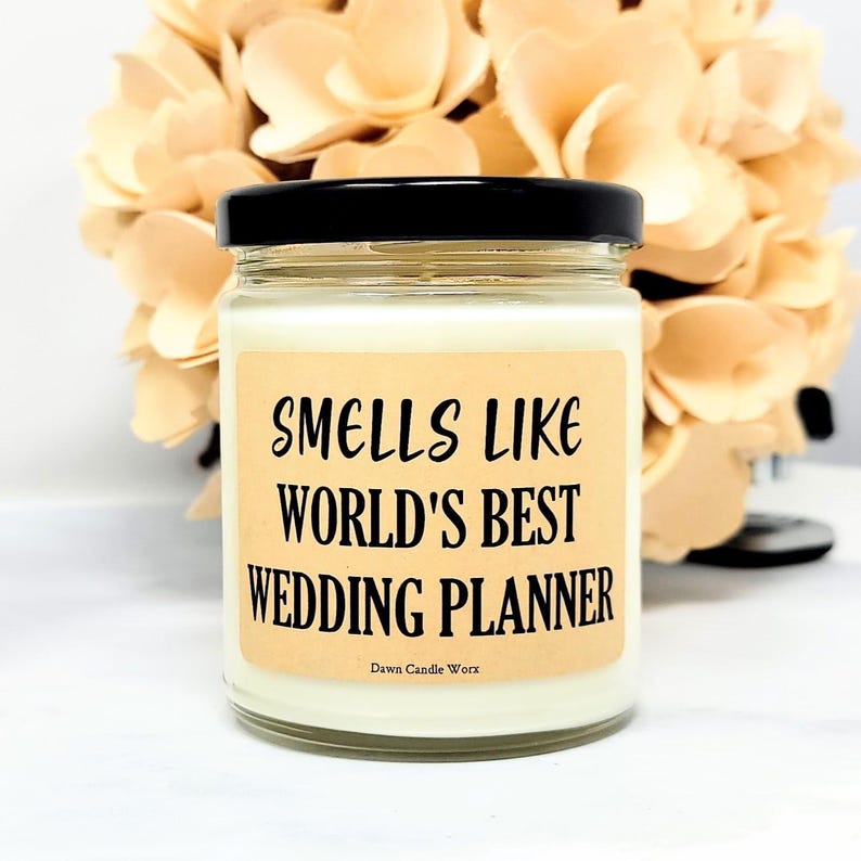 May include: A glass jar candle with a black lid and a label that reads "Smells Like World's Best Wedding Planner." The candle has a light beige label with black text. The background features a bouquet of light-colored flowers.