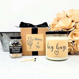 May include: A gift set featuring a candle with "big hugs" text, a jar of matches labeled "The Perfect Match", and a box with a "Thinking of You" label. The candle and match jar have black lids. A black ribbon adorns the gift box.