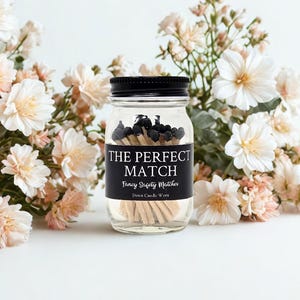 May include: A clear glass jar with a black lid filled with black-tipped matches. The jar has a black label that reads "THE PERFECT MATCH" and "Fancy Safety Matches". The jar is surrounded by white and peach-colored flowers.