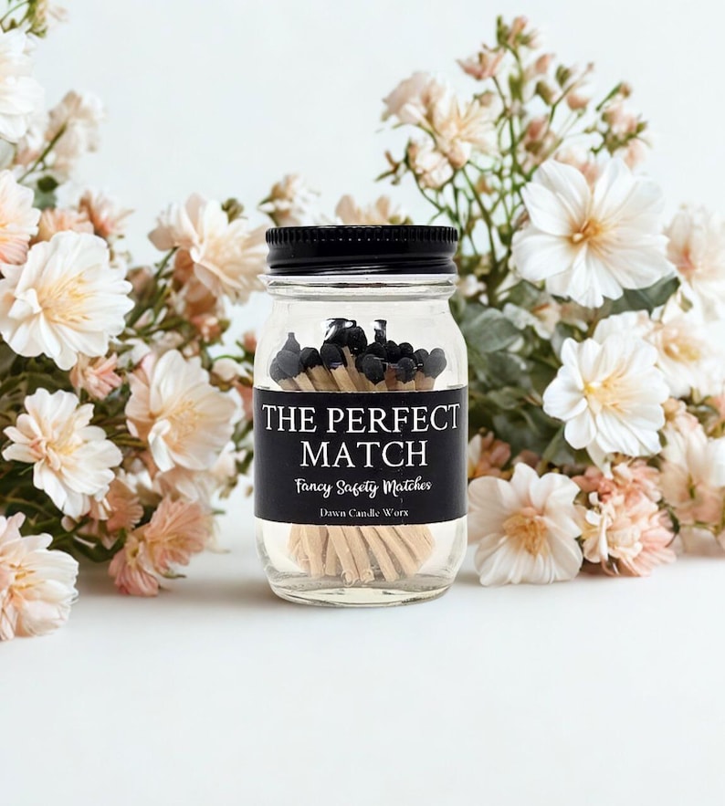 May include: A clear glass jar with a black lid filled with black-tipped matches. The jar has a black label that reads "THE PERFECT MATCH" and "Fancy Safety Matches". The jar is surrounded by white and peach-colored flowers.