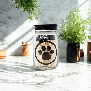 May include: A clear glass mason jar with a black screw-top lid, filled with wooden matches that have black tips. The front of the jar features a black circular graphic containing a solid black paw print design centred on the glass surface.