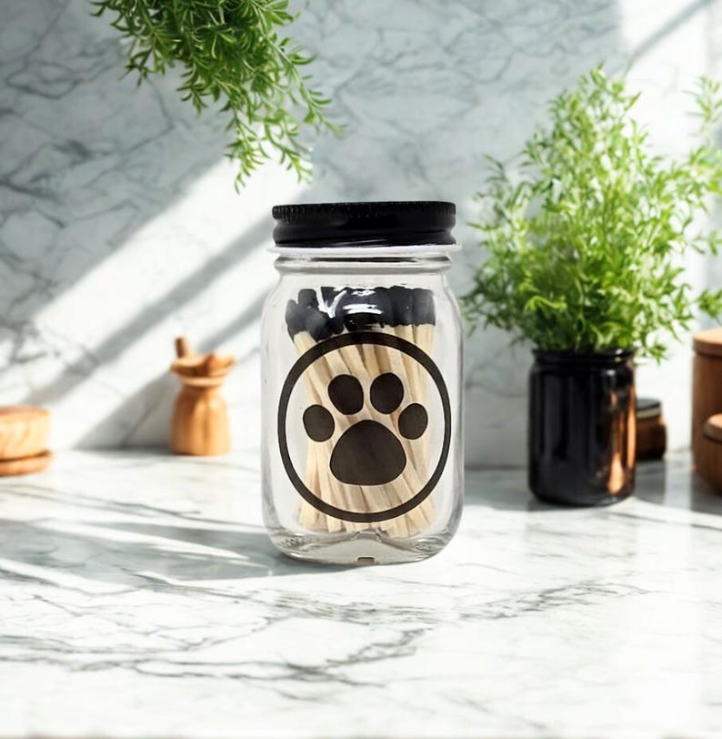 May include: A clear glass mason jar with a black screw-top lid, filled with wooden matches that have black tips. The front of the jar features a black circular graphic containing a solid black paw print design centred on the glass surface.