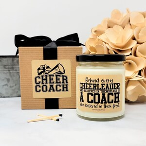 May include: A square cardboard gift box with a black ribbon and a label reading Cheer Coach, next to a glass candle jar with a black lid and a label reading Behind every CHEERLEADER WHO BELIEVES IN THEMSELVES IS A COACH who believed in them first.
