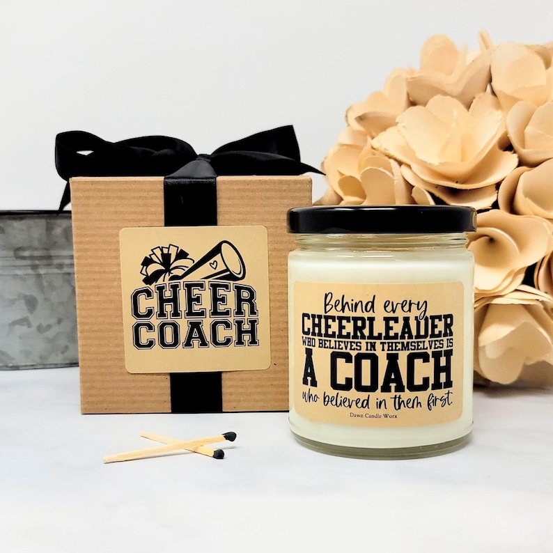 May include: A square cardboard gift box with a black ribbon and a label reading Cheer Coach, next to a glass candle jar with a black lid and a label reading Behind every CHEERLEADER WHO BELIEVES IN THEMSELVES IS A COACH who believed in them first.