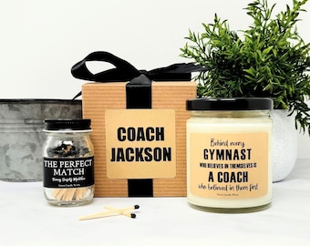 May include: A gift set featuring a brown cardboard box with a black ribbon, a small glass jar of matches labeled THE PERFECT MATCH, and a glass candle jar with a label reading Behind every GYMNAST WHO BELIEVES IN THEMSELVES IS A COACH.