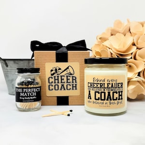 May include: A gift set featuring a square cardboard box with a black ribbon, a small glass jar of matches labeled THE PERFECT MATCH, and a glass candle jar with a label reading Behind every CHEERLEADER WHO BELIEVES IN THEMSELVES IS A COACH.