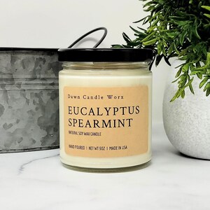 May include: A clear glass jar candle with a black lid and a tan label. The label reads Dawn Candle Worx, Eucalyptus Spearmint, Natural Soy Wax Candle, Hand Poured, Net Wt 9oz, Made in USA. The candle contains off-white wax and sits upright.