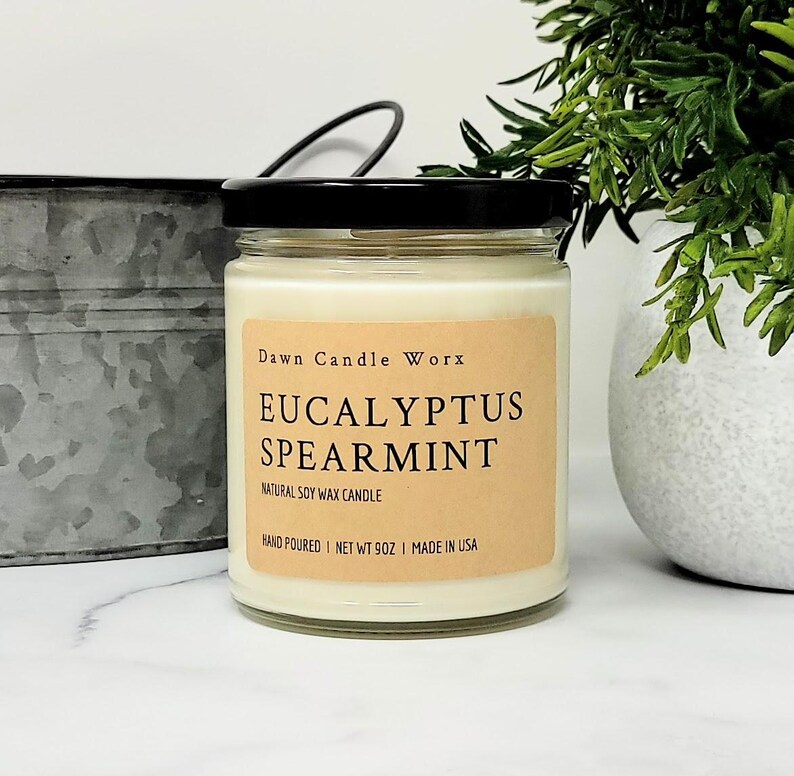 May include: A clear glass jar candle with a black lid and a tan label. The label reads Dawn Candle Worx, Eucalyptus Spearmint, Natural Soy Wax Candle, Hand Poured, Net Wt 9oz, Made in USA. The candle contains off-white wax and sits upright.