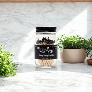 May include: A clear glass jar with a black screw-top lid filled with wooden matches featuring black tips. A black label on the front displays the white text THE PERFECT MATCH, Fancy Safety Matches, and Dawn Candle Worx in a serif font.