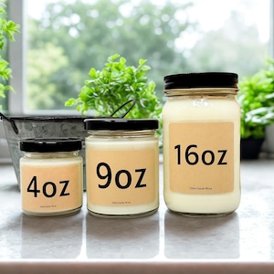 May include: Three glass jars of scented candles with black lids. The jars are labeled 4oz, 9oz, and 16oz. The candles have a creamy white wax and are on a light-colored surface. The background includes greenery and a metal bucket.