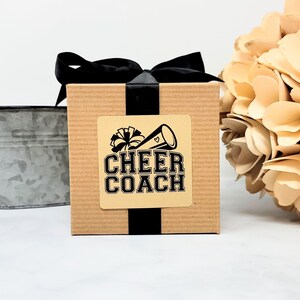 May include: A square, corrugated cardboard gift box wrapped with a black satin ribbon tied in a bow. A square sticker on the front features a black graphic of a megaphone and pom-pom with the text CHEER COACH in a bold, block-style font.