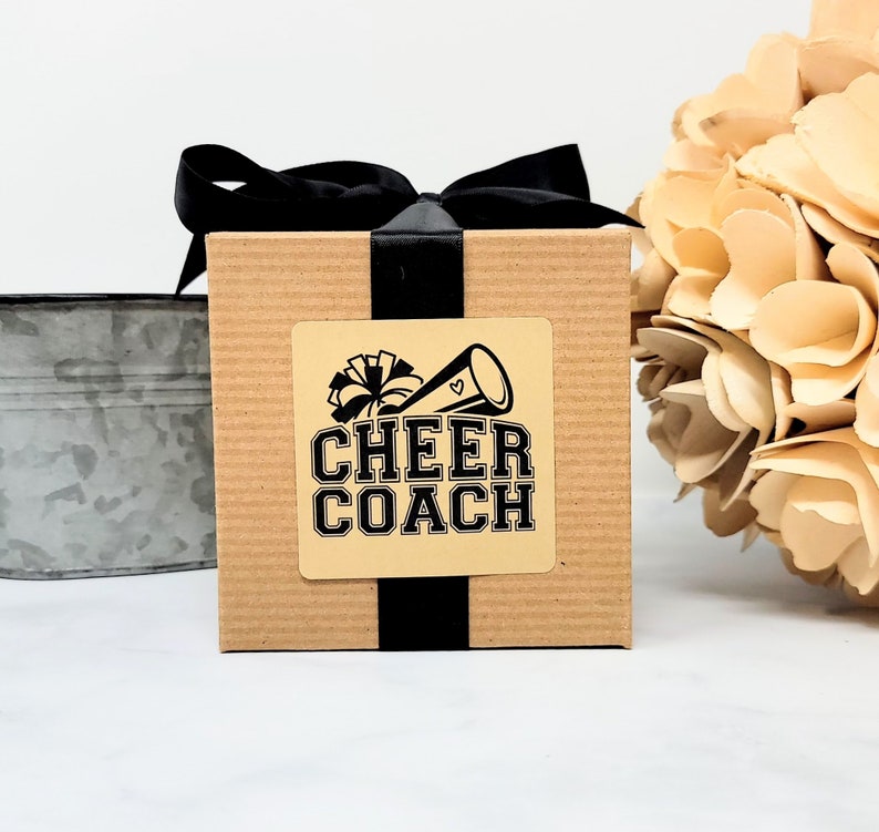 May include: A square, corrugated cardboard gift box wrapped with a black satin ribbon tied in a bow. A square sticker on the front features a black graphic of a megaphone and pom-pom with the text CHEER COACH in a bold, block-style font.