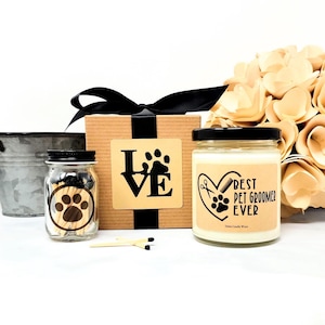 May include: A gift set for a pet groomer, featuring a candle, matches, and a decorative box. The candle has a black lid and a label that reads "Best Pet Groomer Ever." The box has a black ribbon and a paw print design.