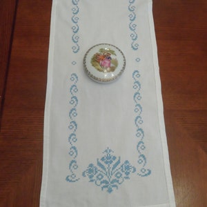 May include: A white rectangular table runner featuring a blue cross-stitch floral border pattern and a small round porcelain trinket box with a colorful illustration of two figures in a landscape setting placed in the center of the runner.