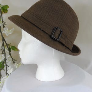May include: A brown fedora-style hat with a structured brim and a dark buckle detail. The hat is displayed on a white mannequin head, showcasing its shape and design. The hat's texture appears to be ribbed.