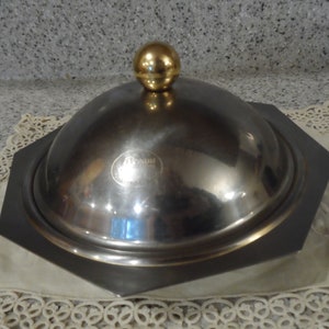May include: A stainless steel serving dish with a gold-colored spherical handle. The dish has a dark gray, octagonal base and a domed lid. The dish is sitting on a cream-colored, lace-edged cloth.