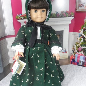 May include: A doll wearing a green dress with white flowers and a black sash. The doll is wearing a green velvet bonnet with white trim. The doll is holding a small red and white gift box.
