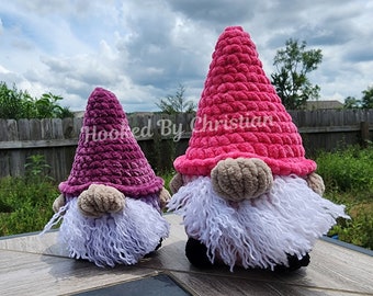Puede incluir: Dos gnomos de crochet, uno rosa y otro morado, con barbas blancas. Los gnomos están sentados en una superficie de madera. El texto "Hooked By Christian" es visible en el fondo.