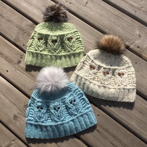 May include: Three crocheted owl hats with faux fur pom poms. The hats are in green, cream, and blue.