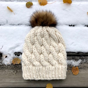 May include: A white knitted beanie with a cable knit pattern and a brown faux fur pom pom.
