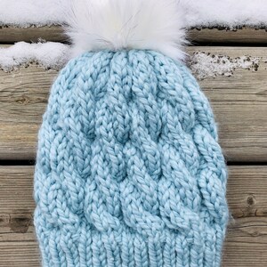 May include: A light blue knitted beanie with a white faux fur pom pom. The beanie has a cable knit pattern.