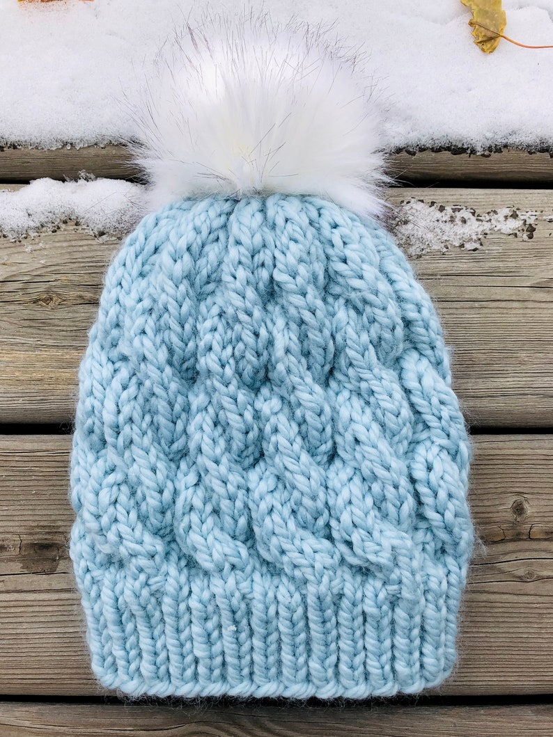 May include: A light blue knitted beanie with a white faux fur pom pom. The beanie has a cable knit pattern.