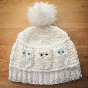 May include: White crocheted beanie with a white pom pom and blue and black buttons in the shape of owls.