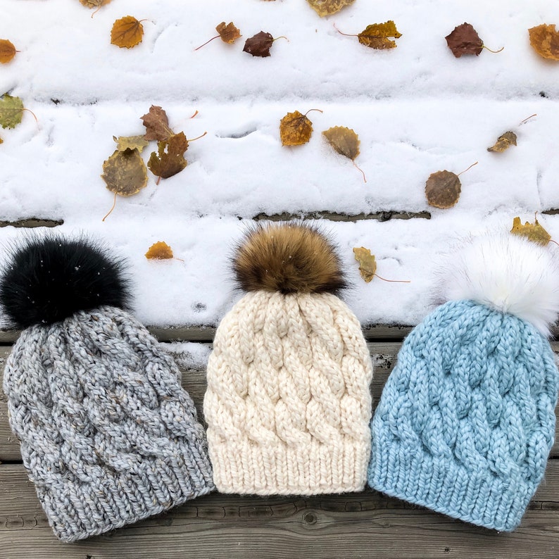 May include: Three knitted hats with pom poms. The hats are in different colours: grey, white, and blue. The hats are sitting on a wooden surface with snow and leaves in the background.