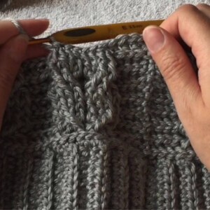May include: Close-up of a person crocheting a gray knitted fabric with a crochet hook. The fabric has a cable knit pattern.