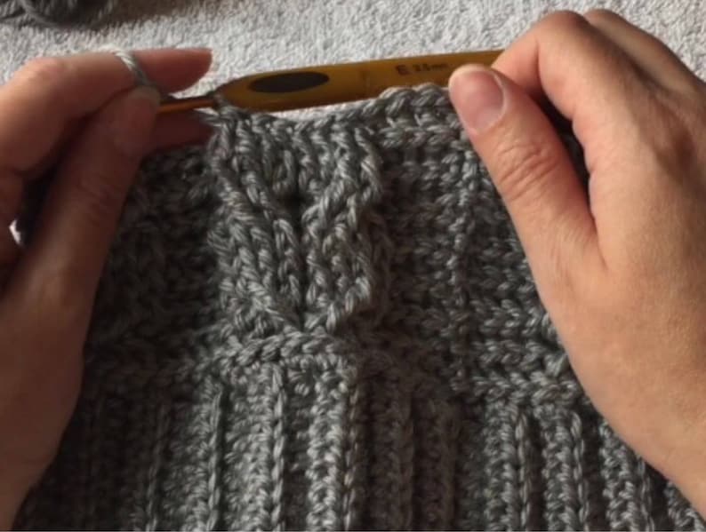 May include: Close-up of a person crocheting a gray knitted fabric with a crochet hook. The fabric has a cable knit pattern.