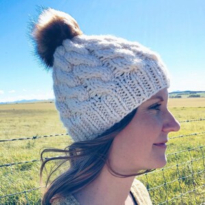 May include: A white knitted beanie with a brown faux fur pom pom. The beanie has a cable knit pattern and is being worn by a person with brown hair.