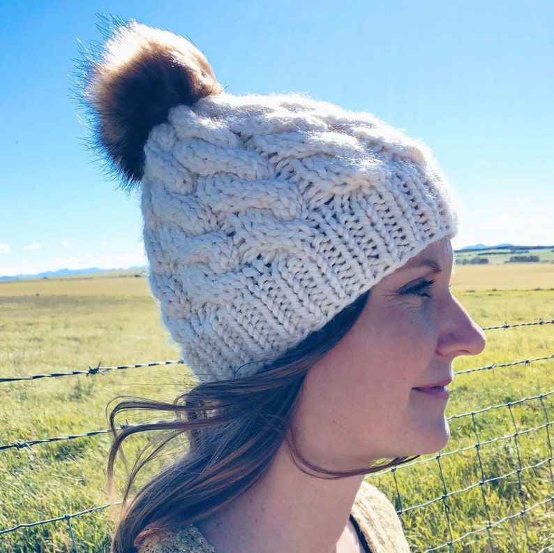 May include: A white knitted beanie with a brown faux fur pom pom. The beanie has a cable knit pattern and is being worn by a person with brown hair.