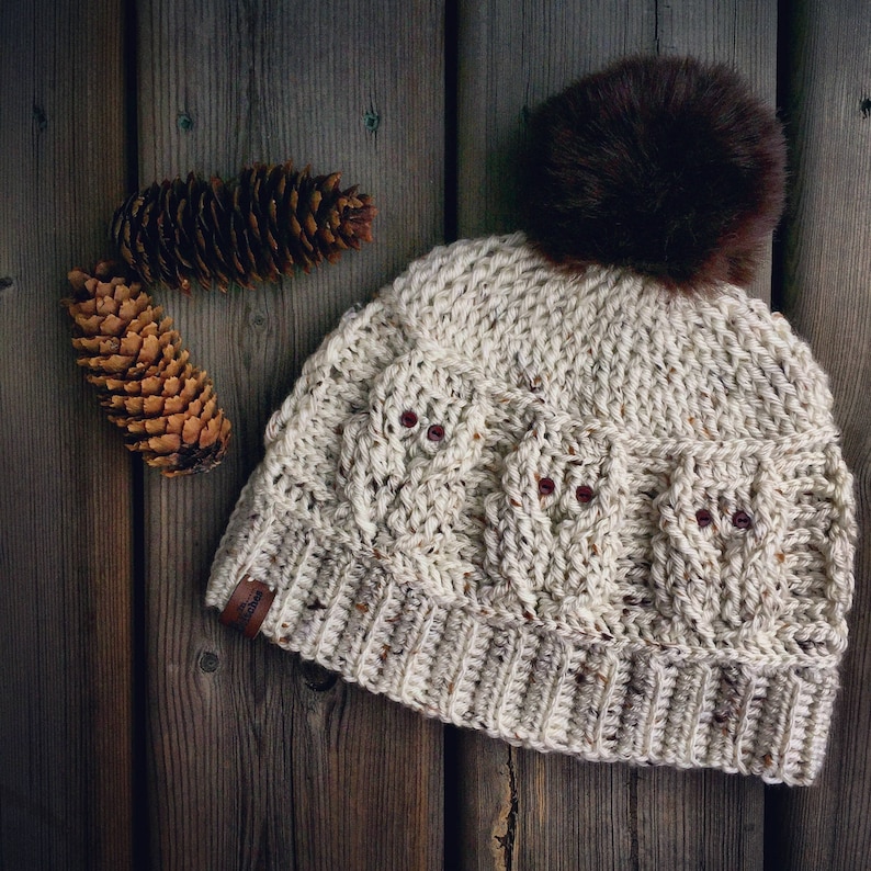 May include: A cream-colored crocheted beanie with a brown faux fur pom pom. The beanie has a cable knit pattern and three red buttons.