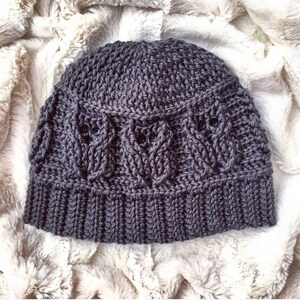 May include: A gray crocheted beanie hat with a textured pattern. The hat has a folded brim and a cable knit design.