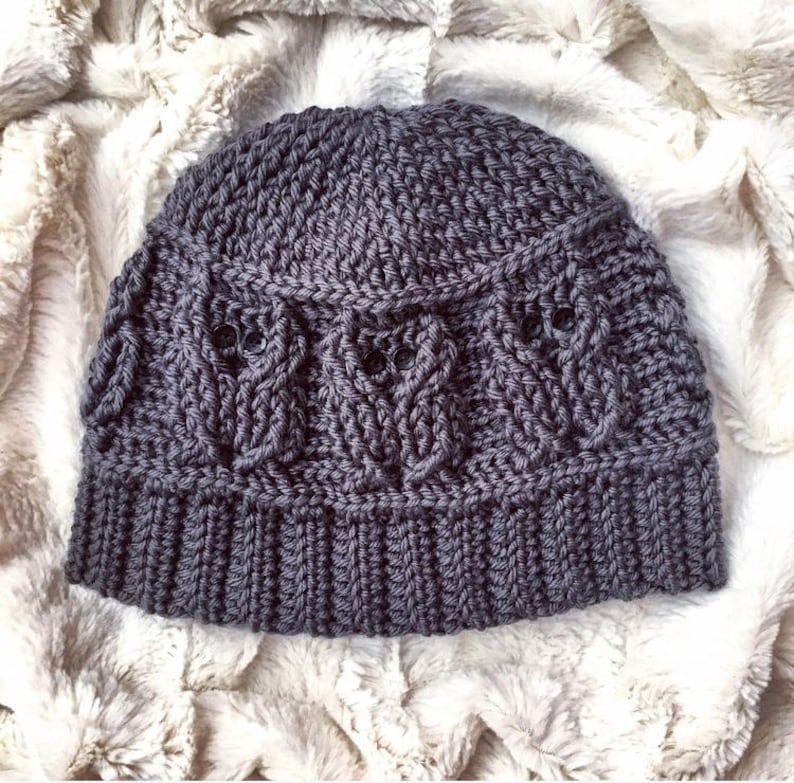 May include: A gray crocheted beanie hat with a textured pattern. The hat has a folded brim and a cable knit design.