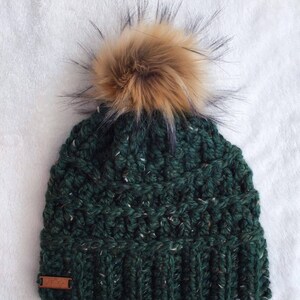 May include: A green knitted beanie with a brown faux fur pom pom. The beanie has a textured knit pattern.