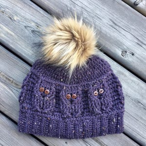 May include: Purple knitted beanie with a brown faux fur pom pom and brown button accents.