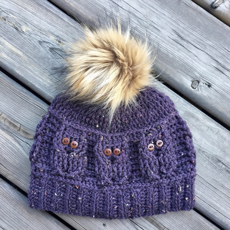 May include: Purple knitted beanie with a brown faux fur pom pom and brown button accents.