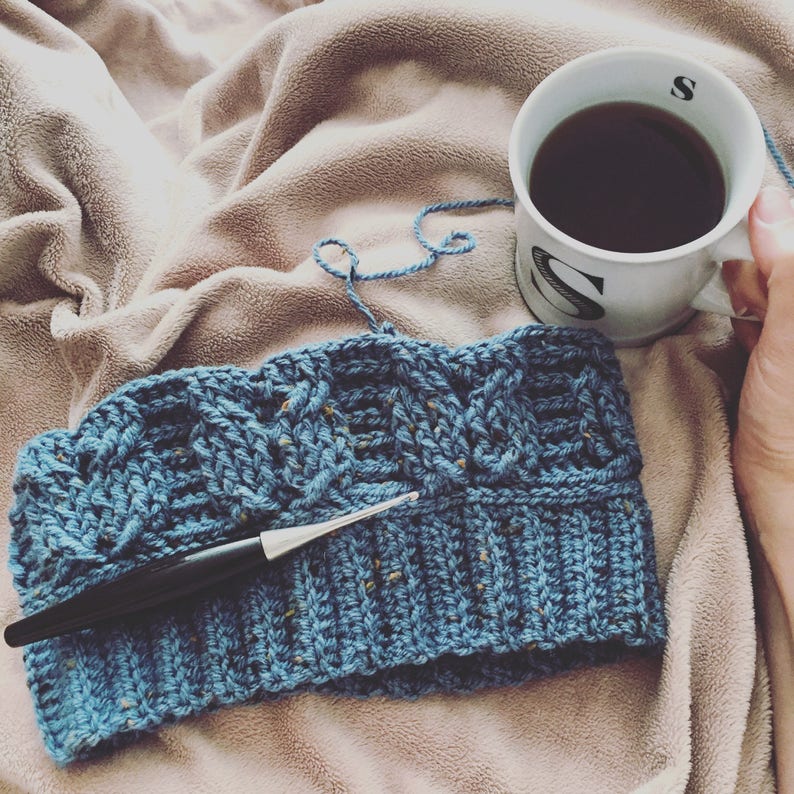 May include: A blue crocheted hat with a cable knit pattern and a crochet hook lying on a beige blanket. A white mug with the letter "S" on it is filled with coffee.
