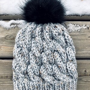 May include: A grey and white cable knit beanie with a black faux fur pom pom. The beanie is made from a soft, warm yarn and is ideal for cold weather.